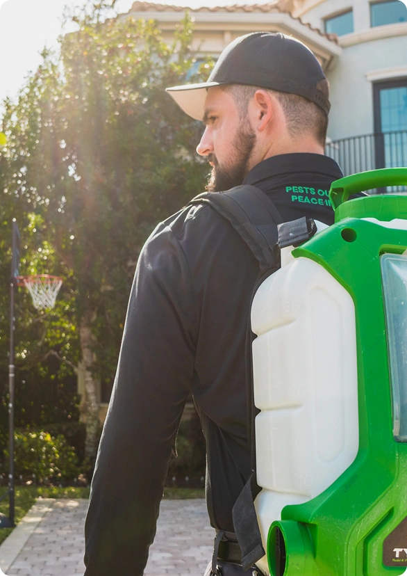 Man wearing a cap and carrying a backpack sprayer, standing outdoors near a house in the service area.