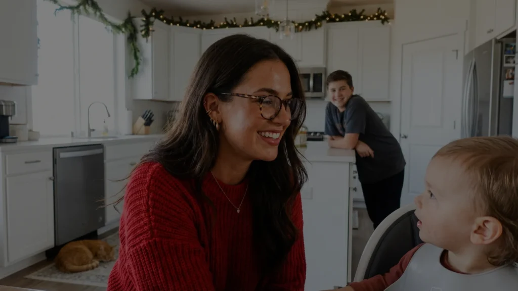 Woman in red sweater smiling at a young child in a kitchen, with another child leaning on the counter in the background.